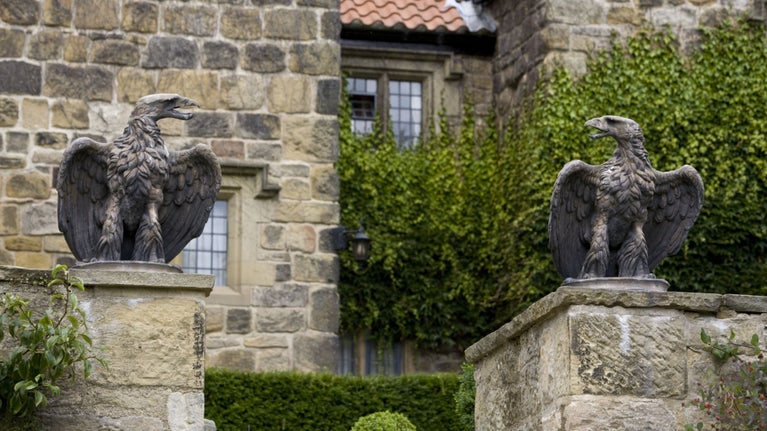 Two stone eagles sitting on top of stone pillars. A large stone building covered in ivy is in the background.